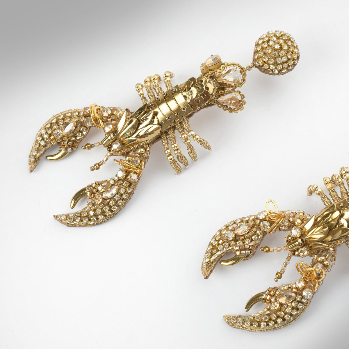 Close up of a pair of gold lobster-shaped drop earrings embellished with small gold stones, shown on a light gray gradient background.