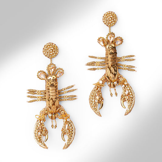 A pair of gold lobster-shaped drop earrings embellished with small gold stones, shown on a light gray gradient background.