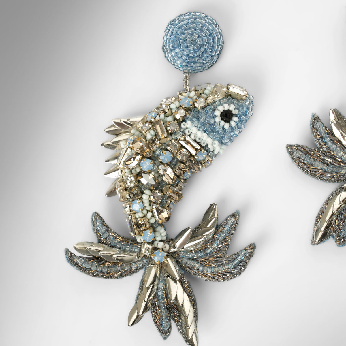 Close up of a pair of ornate fish-shaped earrings embellished with light blue stones, clear crystals, and silver-toned metal, shown on a light gray background.