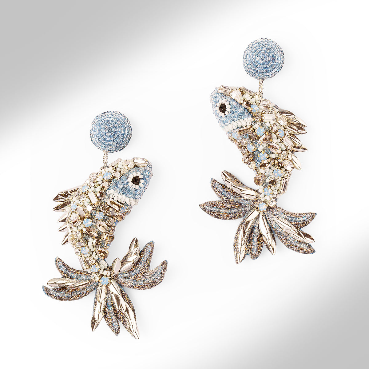 A pair of ornate fish-shaped earrings embellished with light blue stones, clear crystals, and silver-toned metal, shown on a light gray background.