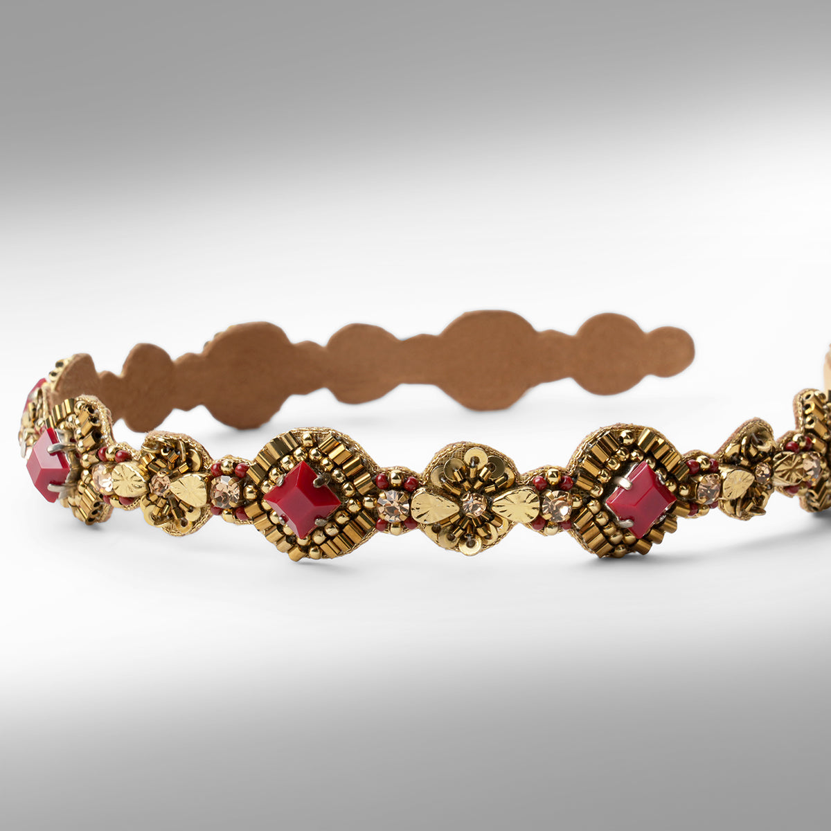 An ornate gold-toned headband with maroon stone accents shown against a neutral gray background.