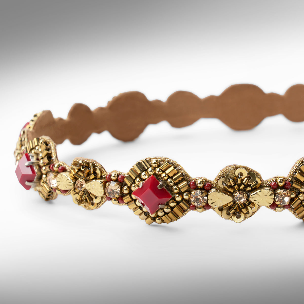 Close up an ornate gold-toned headband with maroon stone accents shown against a neutral gray background.