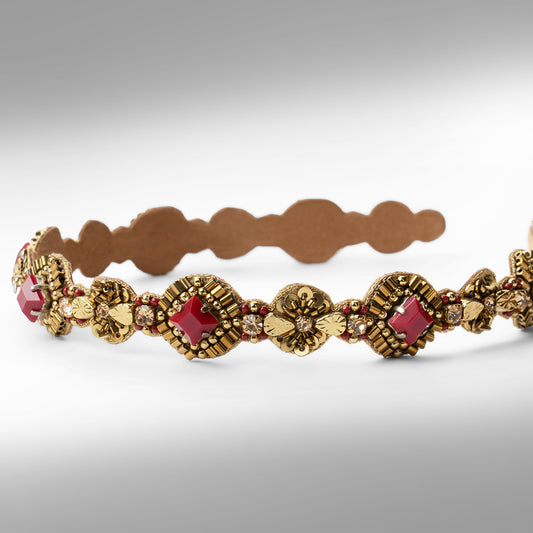 An ornate gold-toned headband with maroon stone accents shown against a neutral gray background.