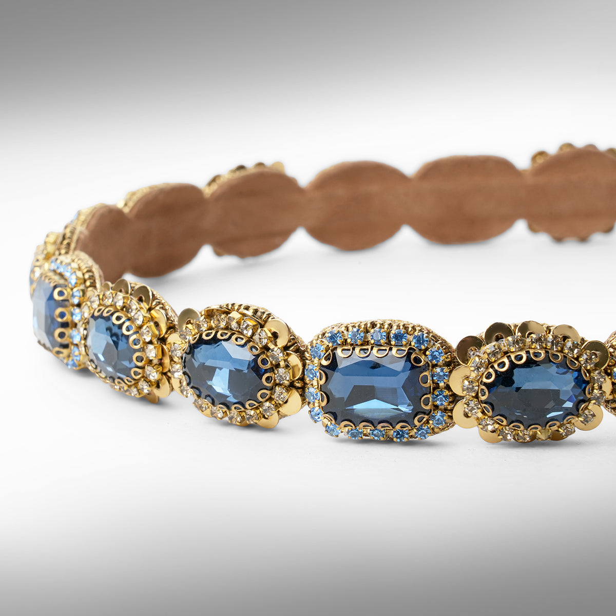 A gold-toned scalloped hard headband featuring oval sapphire stones in ornate settings, displayed on a neutral gray background.