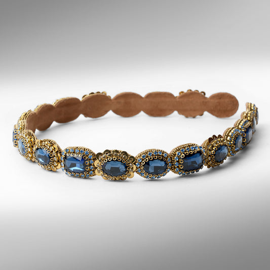 A gold-toned scalloped hard headband featuring oval sapphire stones in ornate settings, displayed on a neutral gray background.