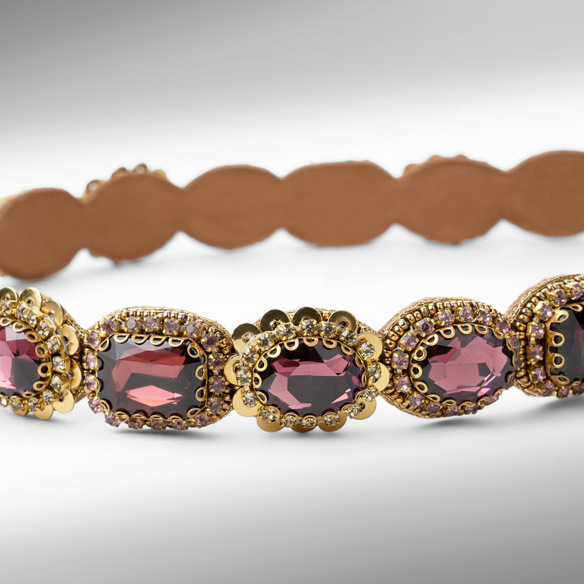 Close up of a gold-toned scalloped hard headband featuring oval burgundy stones in ornate settings, displayed on a neutral gray background.