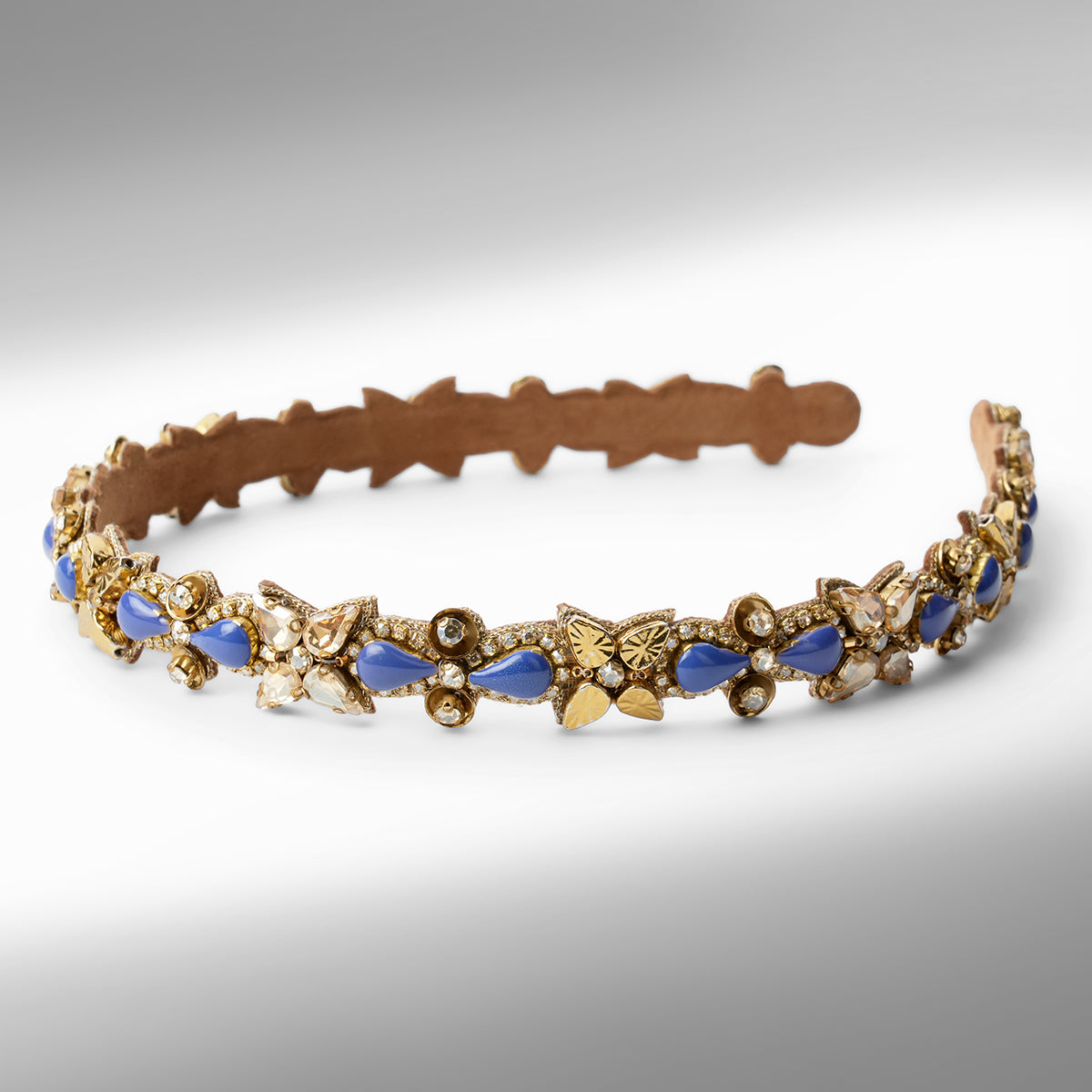 An ornate gold-tone headband decorated with blue and champagne  teardrop-shaped stones displayed on a neutral gray background.