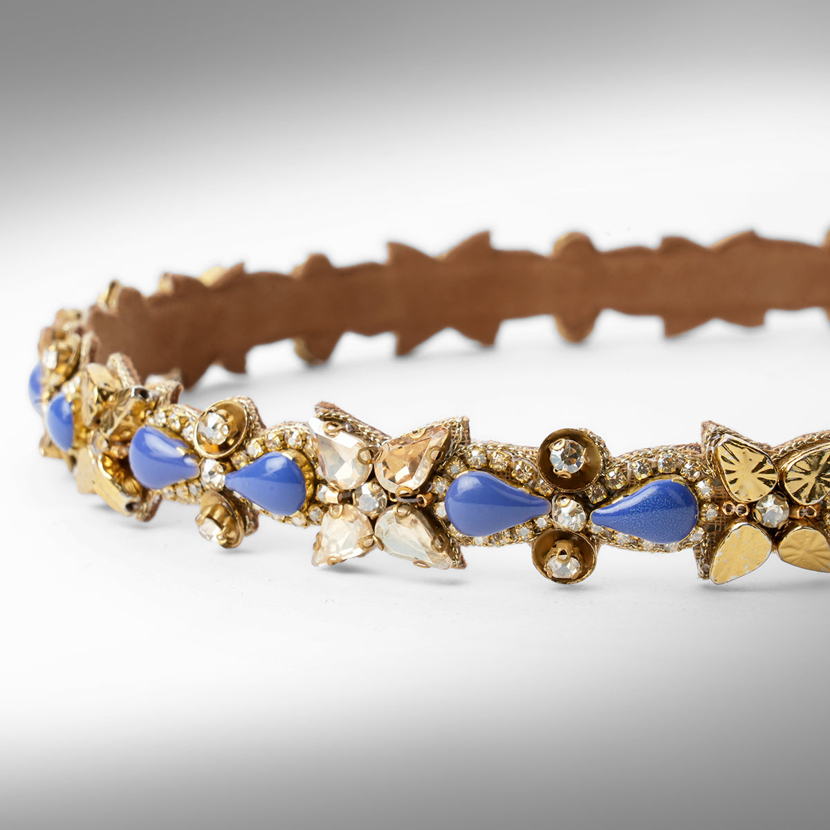 An ornate gold-tone headband decorated with blue and champagne  teardrop-shaped stones displayed on a neutral gray background.