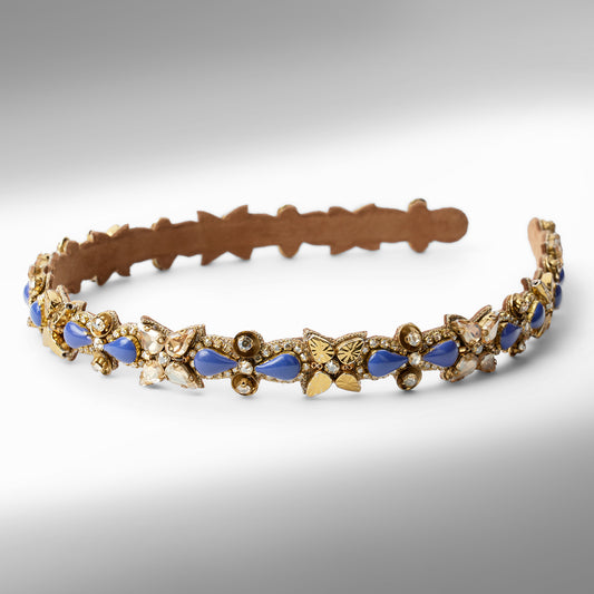 An ornate gold-tone headband decorated with blue and champagne  teardrop-shaped stones displayed on a neutral gray background.