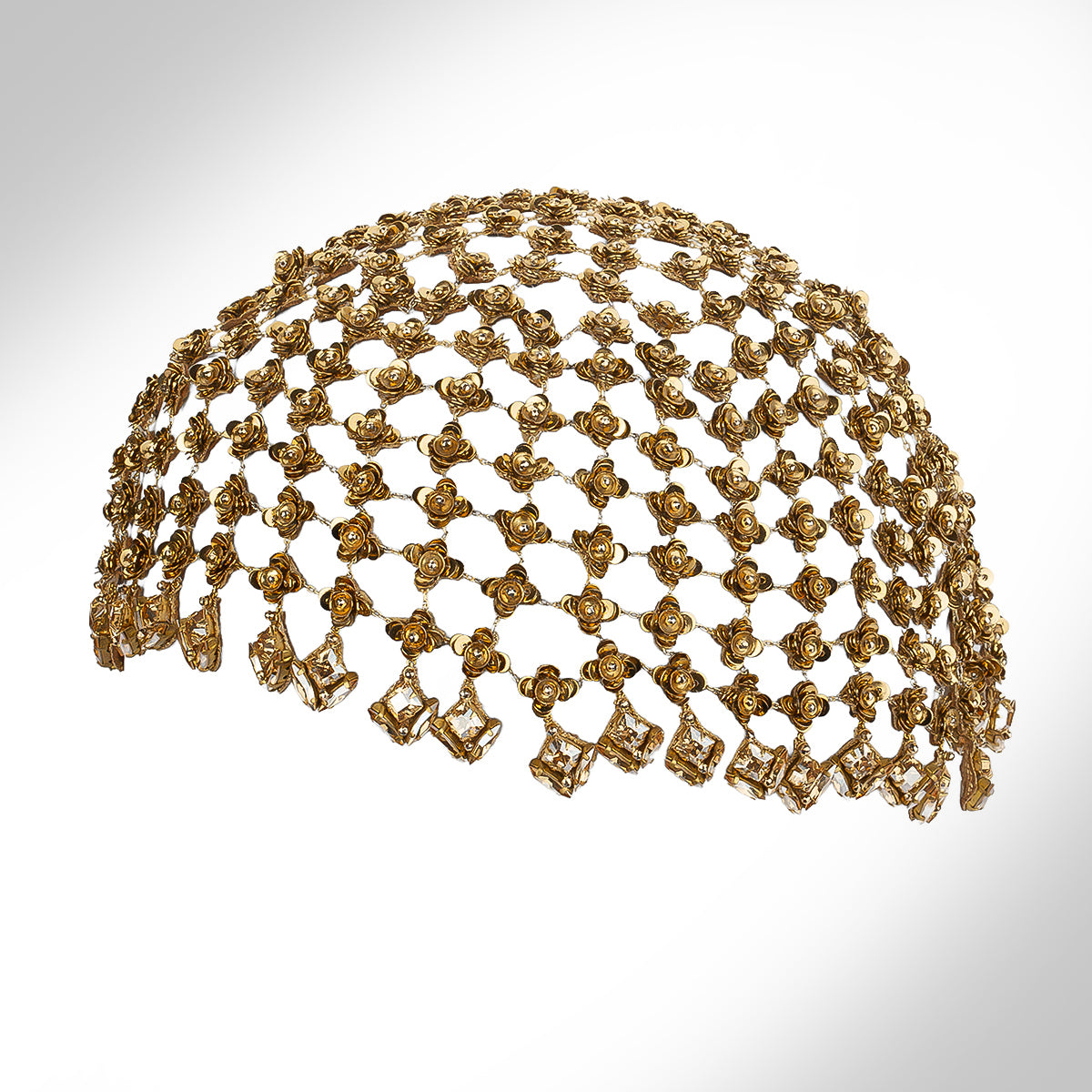 An ornate gold tone headpiece with intricate openwork detailing and dangling floral elements, shown against a light gray gradient background.
