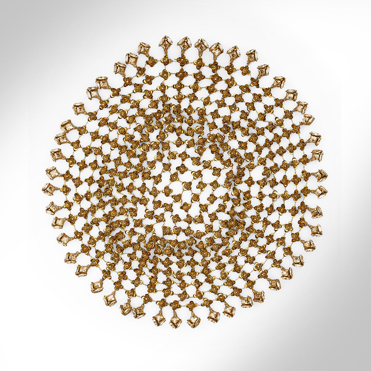 An ornate gold tone headpiece with intricate openwork detailing and dangling floral elements, shown against a light gray gradient background.