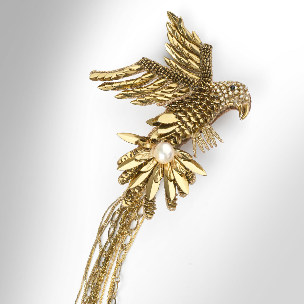 Close up of a gold-toned brooch featuring detailed bird motifs with multiple dangling chain layers, displayed on a grey background.