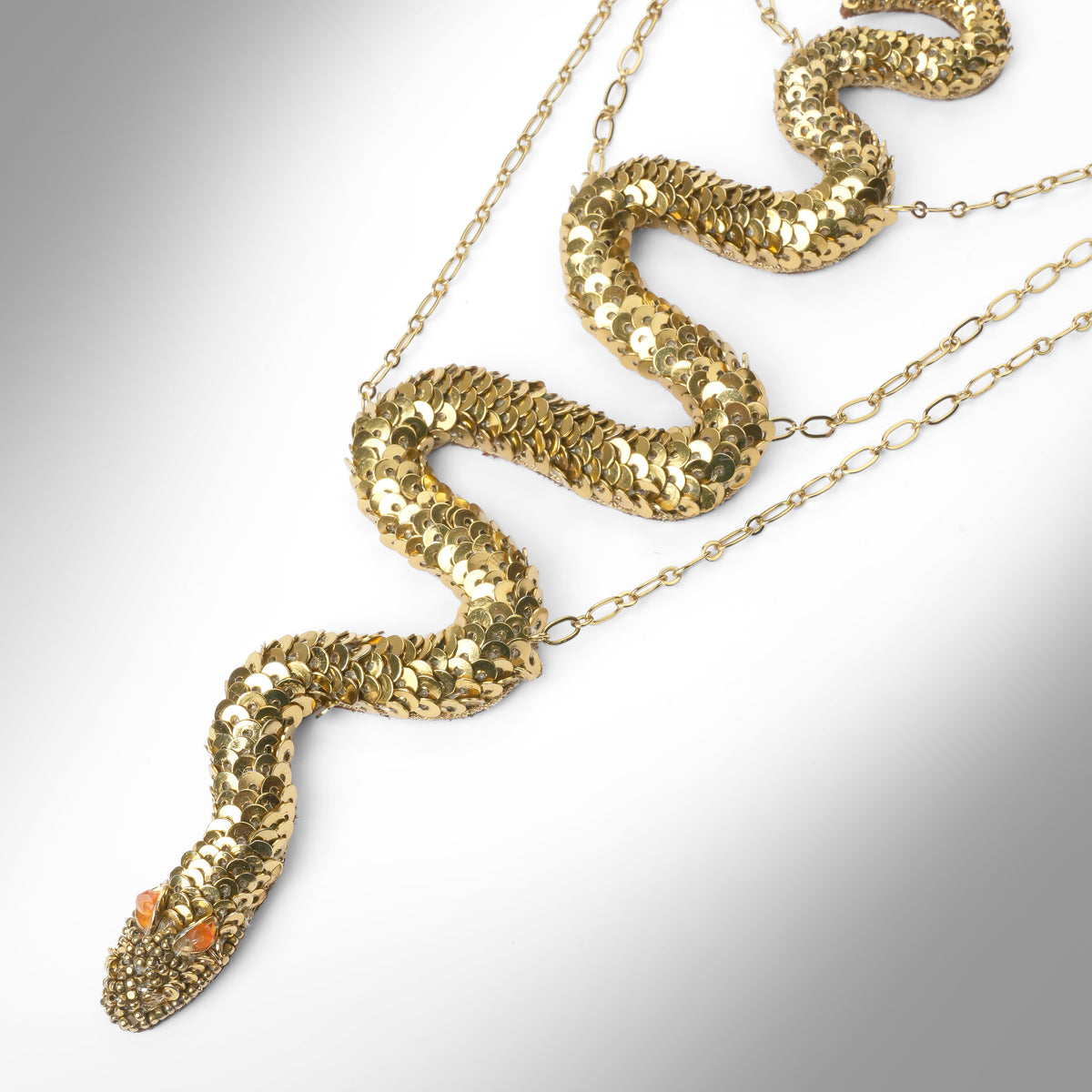 Close up of a gold snake necklace covered in sequins, featuring small orange gemstone eyes, displayed on a light gray gradient background.