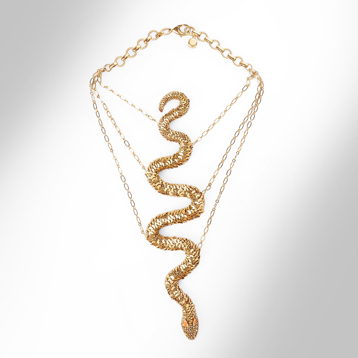 A gold snake necklace covered in sequins, featuring small orange gemstone eyes, displayed on a light gray gradient background.